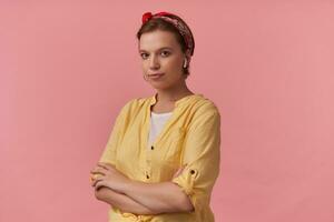 retrato de atractivo joven marrón ojos encantador mujer 20s con natural maquillaje vistiendo blanco camiseta y amarillo camisa y rojo pañuelo posando en contra rosado pared emoción mirando a usted con brazos cruzado foto