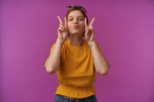 Portrait of european charming woman 20s with natural makeup wearing stylish summer yellow blouse and black bandana with two fingers emotion cheerful flirting eyes closed kiss lips against purple wall photo