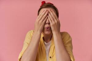 Portrait of european young woman 20s with brown hair with finger over face cover eyes emotion confusion wow hiding playing face with red bandana wearing yellow shirt posing over pink background photo