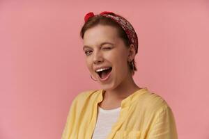 Portrait attractive young brown eyed charming woman 20s with natural makeup wearing white t-shirt and yellow shirt and red bandana posing against pink wall emotion looking at you wink flirt smiling photo