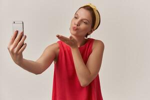 imagen de hermosa joven mujer hacer Disparo Beso emoción vistiendo verano elegante de moda rojo blusa y amarillo pañuelo mirando aparte en inteligente teléfono posando terminado blanco pared con brazos gesticula Beso mosca foto