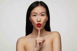 Closeup of attractive young dark haired woman raising forefinger to her red lips while looking at camera, asking to be quiet while standing over white background photo