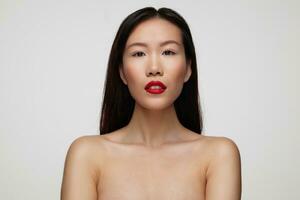 Portrait of beautiful brown-eyed young dark haired woman with red lips looking calmly at camera and keeping her hands down while standing over white background photo