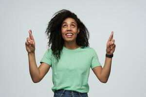 alegre atractivo africano americano joven mujer con largo Rizado pelo en menta camiseta mantiene dedos cruzado y haciendo un deseo aislado terminado blanco antecedentes foto