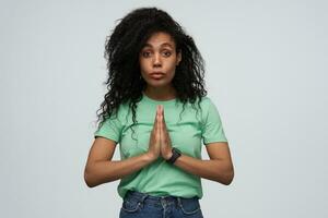 Sad upset african american young woman with long curly hair in mint tshirt keeps hands in praying position and begging isolated over white background photo