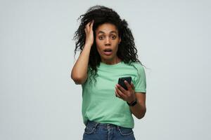 Amazed shocked african american young woman with long curly hair in mint tshirt keeps hands on head and using mobile phone isolated over white background photo