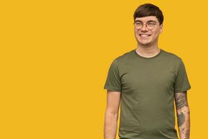Indoor shot of young attractive male, in round stylish glasses and wears green t-shirt, smiles broadly with happy facial expression. isolated over yellow background. photo