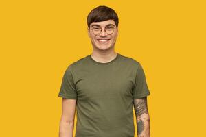 Indoor shot of young attractive male, in round stylish glasses and wears green t-shirt, smiles broadly with happy facial expression. isolated over yellow background. photo