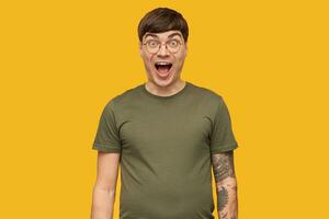 Amazed young male, happy looks into camera with open mouth widely, wears green t-shirt and round glasses. Isolated over yellow background photo