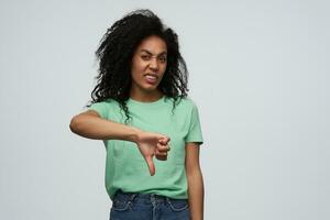 enojado irritado africano americano joven mujer con largo Rizado pelo en menta camiseta mira irritado y demostración pulgares abajo aislado terminado blanco antecedentes foto