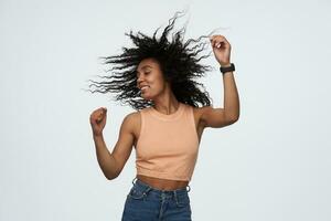 Cheerful joyful african american young woman with eyes closed and flying curly hair dancing and having party isolated over white background photo