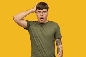 Young shocked male, looking into camera with opened widely eyes and mouth, touching his cheek. Isolated over yellow background. photo