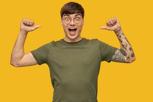 Indoor shot of young happy guy, looking directly into camera with widely opened eyes, point with both thumbs at himself. isolated over yellow background. photo