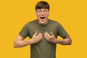 Young happy male, with shocked facial expression, wears green t-shirt, looks directly into camera with smile and opened eyes while receiving a good news, isolated over yellow background. photo