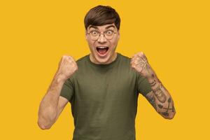 Indoor shot of young male, wears green t-shirt and round stylish spectacles, shouting with widely opened mouth and eyes while watching football game. Isolated over yellow background. photo