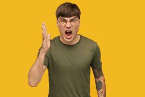 Young angry male student, shouting into camera with negative facial expression, raised his hand. Isolated over yellow background. photo