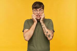 Young stylish man keeps eyes widely opened with shocked, amazed facial expression while watching horror movie, wears green t-shirt and round glasses. Isolated over yellow background photo