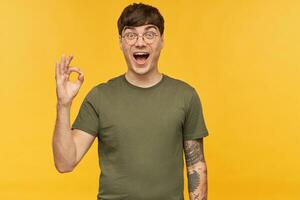 Indoor portrait of young man, shows ok sign with his hand, smiles broadly and looking directly into camera with happy facial expression. Isolated over yellow background. photo