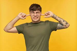 Indoor shot of young male student, looking upwards with negative, tired facial expression, plugs his ears with fingers because a lot of noise. Isolated over yellow background. photo