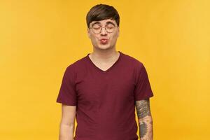 Young flirty male, wears dark red t-shirt and round glasses, keeps his eyes closed while kissing. Isolated over yellow background. photo
