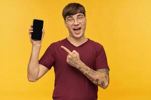 Indoor shot of amazed, shocked young male, wears red t-shirt, pointing with a finger at blank black display of his phone. Isolated over yellow background. photo