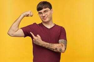 indoor shot of young male student, smiling and pointing with a finger at his biceps, shows how strong he is. Isolated over yellow background. photo