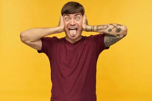 portrait of young funny male, wears red t-shirt, keeps his eyes and mouth widely opened, show his tongue and closed his ears with hands while playing with younger brother. Isolated photo