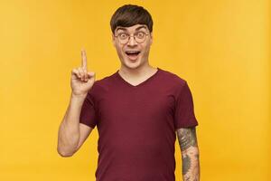 I have an idea. Indoor shot of young student male, smiles broadly and have amazed facial expression, points with a finger upwards. Isolated over yellow background. photo