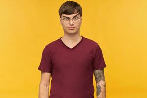 Indoor shot of serious and confused young male, looks directly into camera with irritated facial expression, keeps his eyebrow raised, wears red t-shirt and round glasses. Isolated photo