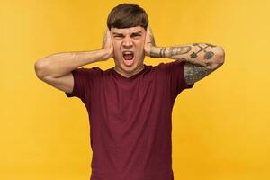 Indoor shot of irritated tired young male student, closed his ears with arms, shouting and want silence. Isolated over yellow background photo