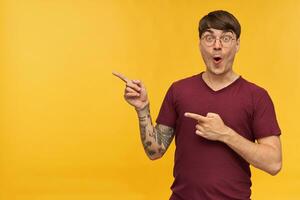Portrait of young male student, wears red t-shirt and round spectacles, pointing with a finger at copy space with amazed, shocked, facial expression. Isolated over yellow studio background. photo