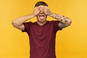Indoor shot of funny young male, closed his eyes with arms, shows tongue while waiting for surprise, isolated over yellow background photo