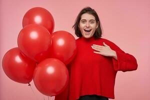 Indoor portrait of young happy positive brunette female, keeps her hand on chest, mouth widely opened while get a gift. Isolated over pink background photo