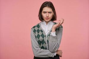 Indoor shot of young brunette student female, starring into camera with angry, mad facial expression, keeps her hand folded. Isolated over pink background. photo