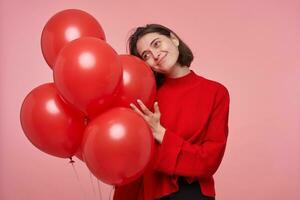 Indoor portrait of young adult brunette female, feels happy with balloons while in party. Isolated over pink background photo