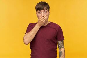 Indoor shot of shocked young male, keeps his eyes widely opened, close his mouth with palm and has amazed facial expression. Wears re t-shirt. Isolated over yellow background. photo
