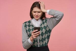 Indoor portrait of young sad unhappy female student, keeps her hand on head while reading bad news . Isolated over pink background photo