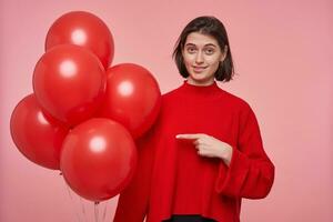 Indoor shot of young female wears red sweater indicates with finger at red bright balloons. Isolated over pink background photo