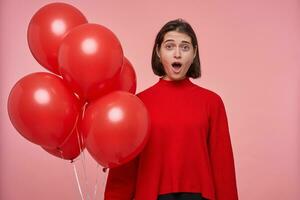 Young brunette female wears bright red sweater keeps mouth widely opened with shocked, surprised facial expression. Isolated over pink background photo