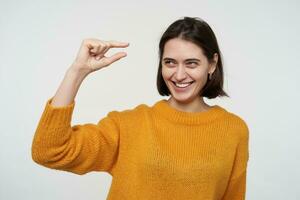 Indoor shot of young positive female wears yellow sweater, showing small gesture, smiles. Isolated over white background photo