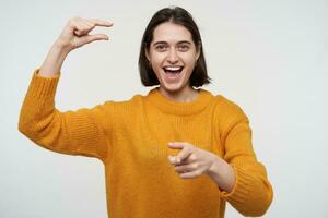 Indoor shot of young positive female wears yellow sweater, showing small gesture, smiles. Isolated over white background photo