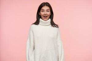 Surprised young lovely brunette female with loose hair looking excitedly at camera with opened mouth and keeping hands along body while posing over pink background photo