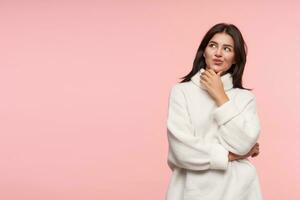 Glad young attractive brown haired female with natural makeup pursing her lips while looking pensively upwards, posing over pink background in white poloneck photo