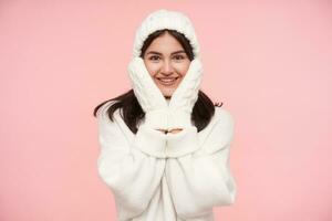 alegre joven de ojos marrones morena hembra vestido en blanco lana acogedor ropa participación su cara con elevado manos y sonriente felizmente a cámara, aislado terminado rosado antecedentes foto