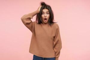 Indoor photo of shocked young brown haired woman raising amazedly her eyebrows while looking dazedly at camera, standing over pink background in casual wear