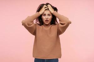 Bemused young bewildered brunette female with natural makeup holding her head with raised hands while looking confusedly at camera, isolated over pink background photo