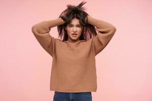 Indoor photo of young pretty brown haired woman cluthing her head with raised hands and looking attentively at camera, isolated over pink background