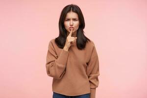 Severe young pretty brunette lady with casual hairstyle frowning her eyebrows while looking seriously at camera and holding index finger on her lips, isolated over pink background photo