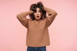 Shocked young pretty brunette lady keeping her hands raised while rumpling her hair and looking surprisedly at camera with wide eyes opened, posing over pink background photo