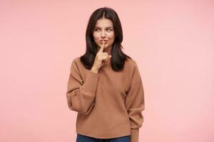 Glad young attractive brown haired woman with loose hair smiling cunningly while raising hand with hush gesture, standing over pink background in casual clothes photo